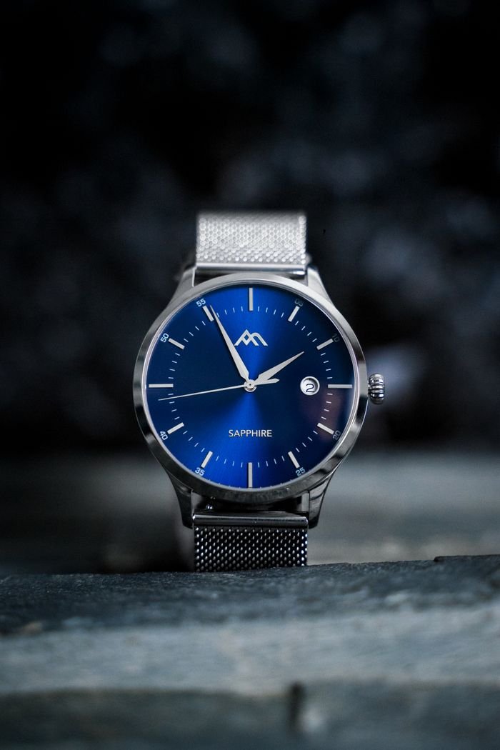 our-mission-01 Close-up shot of an elegant wristwatch with a blue sapphire dial and metal mesh strap.